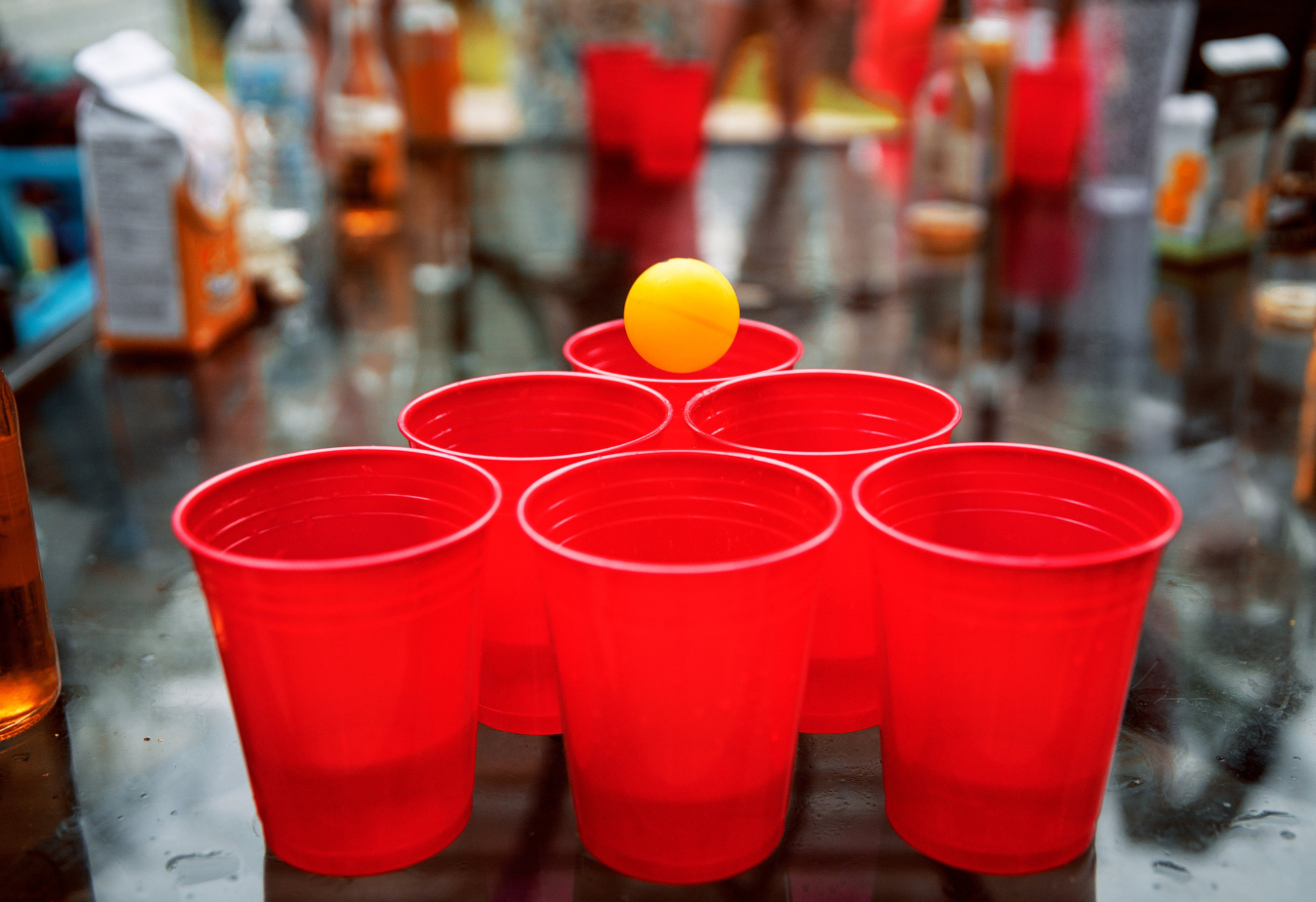 6 red cups in a triangle with a ping pong ball above one cup, alcohol bottles in the background blurred